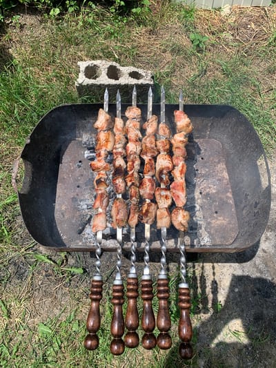 The image shows skewers of raw meat cooking over coals in a black metal barbecue grill. The grill is situated outdoors on a grassy area. There are several cinder blocks to the left of the grill and a concrete surface to the right. The skewers have decorative wooden handles. The meat is cut into uniform cubes and appears to be marinating. The sun is shining, casting shadows on the ground.