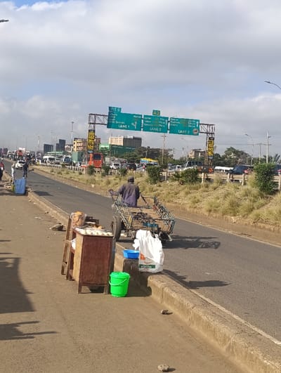 A street view shows a man pulling a cart on a highway shoulder next to a makeshift food stand. The stand has a wooden cabinet, topped with items wrapped in plastic, a blue bucket, and a large white sack. Overhead highway signs indicate directions, including "Exit 9" to Thika and Barnicke in Lane 1, and A2 towards Nairobi, Eldoret, and Kakamega in Lanes 2 and 3. Heavy traffic fills the highway, with numerous cars, trucks, and buses. A grassy area separates the highway lanes.