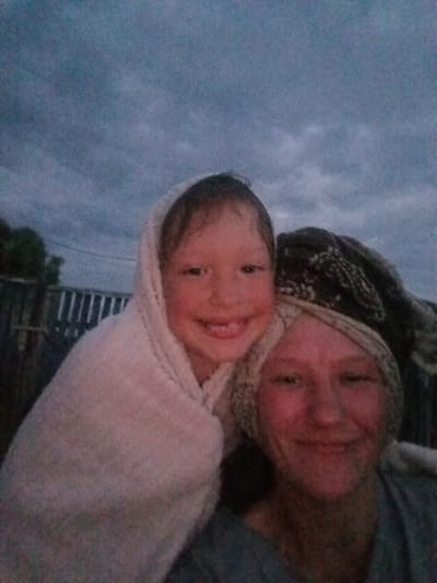 This media file shows a close-up selfie of two people outdoors, likely during twilight. The person in the foreground, on the right, is an adult with reddish skin and a patterned headscarf. They are looking towards the camera with a slight smile and their eyes are partially closed. To the left of the adult is a child, who is wrapped in a light-colored towel and also looking at the camera, smiling broadly. The background suggests a rural setting with a wooden fence and some trees visible. The sky is cloudy and a deep blue, indicating either dusk or overcast conditions. The overall mood appears to be warm and affectionate, with the two individuals sharing a moment together. The image quality is somewhat grainy, typical of low-light photography. No text is visible in the frame.