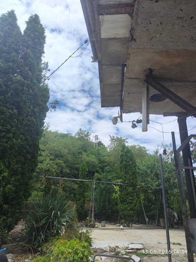 The image depicts an outdoor scene in what appears to be a yard or a semi-rural area, possibly in Dobrotino, Bulgaria. In the foreground, to the left, are several tall evergreen trees. Adjacent to them is a large, spiky plant, likely a yucca, surrounded by smaller green plants and some rusty metal garden decorations. The ground is a mix of dirt and scattered rocks.

In the midground, more trees and foliage create a lush backdrop. A blue pipe runs horizontally across the scene, supported by posts. A black and white soccer ball rests on the ground. To the far right, a metal gate or fence is partially visible.

In the upper portion of the image, a concrete structure, possibly an overhang or roof, is visible, showing signs of wear and some rusting metal supports. Attached to this structure are two security cameras and a long, light-colored fixture that appears to be some kind of light or antenna. Electrical wires are strung across the sky.

The sky is partly cloudy with blue sky visible between white clouds. The timestamp "13.06.2026 14:39" is superimposed on the bottom right corner of the image. No people are present in the scene. The overall impression is one of a somewhat unkempt outdoor space with security features installed.