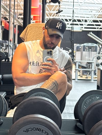 A man with a beard and a black baseball cap with a white logo is taking a mirror selfie in a gym. He is wearing a white sleeveless shirt and black shorts. He has tattoos on his right arm and leg. In front of him, there are several dumbbells. In the background, gym equipment is visible. The image appears to have been taken inside a gym in Emmen, Netherlands.