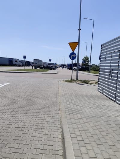 The image depicts an outdoor scene at a parking lot or rest stop, likely in Chomentowo, Poland, given the context. The foreground is dominated by a paved area with interlocking gray concrete pavers. A curb separates this paved area from a strip of grass.
In the mid-ground, there is a road or driveway, and beyond that, a parking area filled with several large trucks and some passenger cars. The trucks are mostly white and gray, with some bearing company logos. One truck has a dark grey trailer with a circular logo. A few people are visible near the trucks, one in a white shirt and dark pants standing near a black car. Another person, wearing a light-colored shirt and dark pants, is walking away from the viewer towards the parked vehicles.
Several signs are present. A blue sign with a white "P" indicates parking. Another sign, a blue circular sign with a white arrow pointing right and upwards, accompanied by a yellow triangular sign above it, signifies a mandatory direction or turn. Streetlights are visible, including one with a security camera attached to it.
In the background, more trucks and vehicles are parked, and a large, corrugated metal building or structure, possibly a warehouse or storage unit, is prominently featured on the right side of the image, its silver surface reflecting the bright daylight. The sky above is clear and bright blue, indicating a sunny day. The overall impression is one of a functional, well-lit service area.