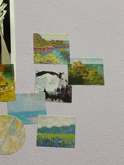 The image displays a collection of small, postcard-like prints affixed to a textured, light purple wall. The prints are arranged in a somewhat scattered pattern, with some overlapping.

One print at the top depicts a rocky coastline with lush green foliage and blue water. To its right, another print shows a small, tan house situated on a grassy hill overlooking a vibrant green sea.

Below these, a print features a black and white scene with a bridge arching over water, and silhouetted figures walking along a waterfront.

To the left of the bridge scene, a vertical print shows a garden path with trees and flowers in warm autumn colors, bordered by a white fence.

Below the garden print, a rectangular print displays a soft blue sky with hints of pink and orange near the horizon, suggesting a sunset or sunrise.

Further down and to the right, a print portrays a landscape with a field of green and blue wildflowers in the foreground and a dense green forest in the background, under a pale sky.

To the bottom left, a circular print features abstract swirls of yellow, white, and light blue, resembling a watercolor or marbling effect.

The prints are diverse in subject matter, ranging from landscapes and seascapes to architectural elements and abstract art. The overall impression is that of a curated collage or personal display on a wall. The location in the city of Jrarat, Armenia, is not indicated by any visual cues within the image.