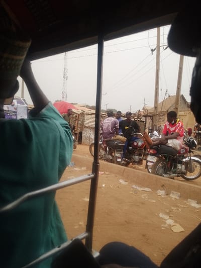 The image depicts a street scene in Lapai, Nigeria, viewed from inside a vehicle, likely a tricycle. In the immediate foreground, the back of a person wearing a green garment and a patterned cap is visible, alongside the vehicle's metal frame. The midground shows a dusty, unpaved roadside with a low concrete curb. At least five men are present, some seated on motorcycles, others standing. One man on the right wears a red and white patterned shirt, white shorts, and a black face mask, sitting on a red motorcycle with his bare feet on the handlebars. Another man in the center wears a black Nike t-shirt and cap, seated on a red motorcycle. Litter, primarily white paper/plastic fragments, is scattered on the ground. In the background, multiple makeshift structures with corrugated metal roofs line the road, interspersed with wooden utility poles carrying overhead wires. A tall metal lattice telecommunications tower is visible against an overcast, light grey sky. To the left, a person holds a red umbrella near some stalls, and part of a light-colored vehicle is visible further back.