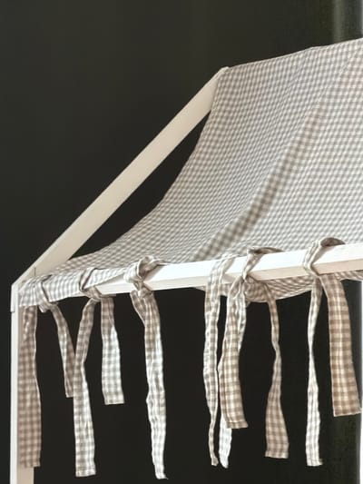 A close-up, medium shot captures a white, tent-like structure with a checkered fabric roof and decorative ties hanging from its edge. The structure appears to be made of white painted wood or metal poles. The fabric is a light gray and white gingham pattern. The background is a solid, dark gray or black color, creating a stark contrast with the lighter foreground elements. The image is tightly framed, focusing on the textures and patterns of the fabric and the structure. There are no people, animals, or explicit signs of location within the city of Magdeburg, Germany.