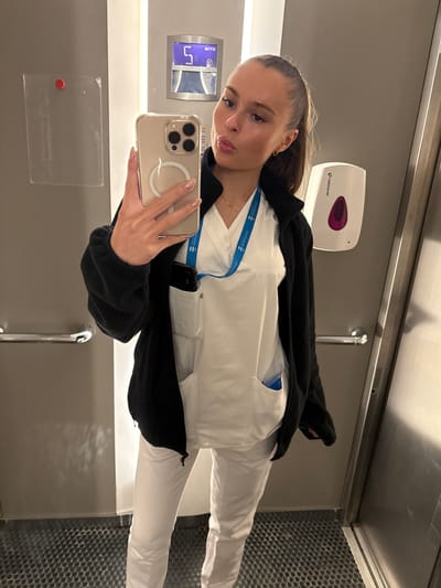A young woman with blonde hair pulled back into a ponytail, wearing white scrubs and a black zip-up jacket, takes a selfie in an elevator. She is holding a gold iPhone in her right hand. A blue lanyard with a card is visible around her neck. A white dispenser is mounted on the wall to her right, and a digital display showing the number "5" is visible on the wall behind her. The elevator has a metal handle on the left wall and a textured metal floor. The overall environment appears to be an indoor, possibly institutional setting like a hospital or healthcare facility in Harstad, Norway.