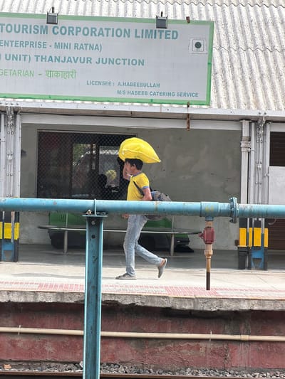 This image, taken in Thanjavur, India, depicts a person carrying a bright yellow bag over their shoulder, holding it with a yellow umbrella to shield themselves from the elements. The individual is wearing a yellow t-shirt and light-colored jeans. They are positioned in front of a building with a sign that reads "TOURISM CORPORATION LIMITED ENTERPRISE - MINI RATNA) UNIT) THANJAVUR JUNCTION VEG CETARIAN - शाकाहारी LICENSEE: A.HABEEBULLAH M/S HABEEB CATERING SERVICE". The sign indicates a food establishment, specifically a vegetarian one. The setting appears to be outdoors, possibly a street or platform, with a textured wall and a blue pipe structure prominent in the foreground. The lighting suggests it is daytime, and the presence of the umbrella hints at either sun or rain. The overall scene captures a momentary pause in the day, with the individual navigating their environment with their load.