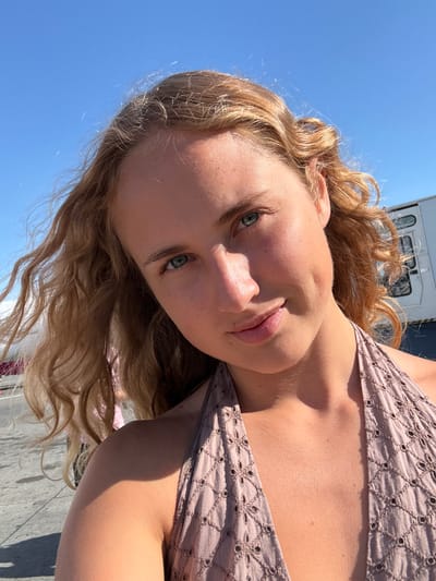 A young woman with wavy blonde hair and blue eyes takes a selfie outdoors under a clear blue sky. She is wearing a light-colored halter top with a textured pattern. Her left cheek has a dimple. The background includes a white trailer or building with windows and a shadowed area. The ground appears to be paved. Based on the provided information, this scene is not set in Kruja, Albania, but rather appears to be an outdoor setting with a modern, possibly industrial or transportation-related backdrop.