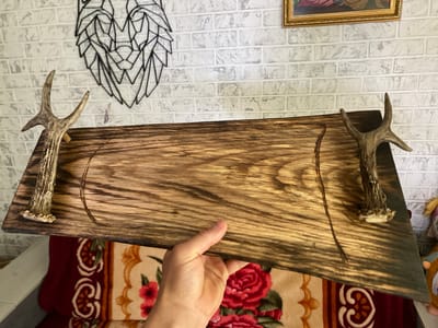 In Negrești, Moldova, a person's hand holds a rustic wooden serving tray adorned with two deer antlers, one on each side, serving as handles. The tray is rectangular with rounded corners and features two subtly carved, curved indentations in the center, suggesting a design for holding food or drinks. The wood has a dark, weathered finish with prominent grain patterns. The background reveals a white brick wall, likely indoors, with a decorative geometric wire art piece of a wolf's head and a framed picture with a gold border hanging above. Below the tray, a richly patterned blanket with floral motifs in red, cream, and maroon is visible, hinting at a cozy interior setting. The lighting appears to be ambient, suggesting daytime, but the specific time is not discernible. The overall impression is one of handmade craftsmanship and a connection to nature, possibly a decorative piece for a home.