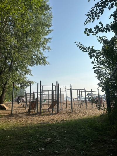 A low-angle shot captures a sandy outdoor gym area with various workout equipment, including pull-up bars and parallel bars, on a beach. The ground is a mix of sand and grass, with trees framing the scene. In the background, the beach is populated with people relaxing under umbrellas and near the water. The sky is clear and blue, suggesting a warm day. This location is identified as the beach at Ust-Kachka, Russia.
