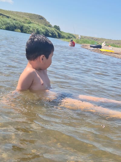 The image depicts a person submerged in shallow, murky water. The subject, a young boy with dark, wet hair, is shown from the chest up, facing away from the camera and looking towards the left. His skin appears to be tanned. The water is rippled, indicating movement or a gentle current, and the bottom is visible in places, revealing rocks or pebbles.
In the background, the shore of a body of water is lined with lush green vegetation that climbs up a gentle slope. The sky above is clear and blue, suggesting a sunny day. Further in the distance, other people can be seen enjoying the water or the shore. One person is kneeling in the water with a fishing rod, while others are present near inflatables and what appears to be a yellow kayak or raft on the shore. The overall scene suggests a recreational outing in a natural environment, likely during warm weather. The image is oriented vertically.