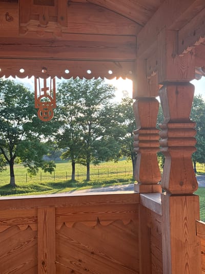 The image shows a close-up view of a wooden porch with a view of trees and a grassy field beyond. The porch features intricate wooden carvings and turned posts. In the foreground, the detailed grain of the wood is visible, highlighting its natural beauty. A decorative wooden element, possibly a religious symbol, hangs from the porch ceiling. The background reveals a bright, sunlit scene with lush green trees and a fence bordering the field. The setting appears to be rural, with the natural landscape dominating the view. There are no people or animals visible in the image.