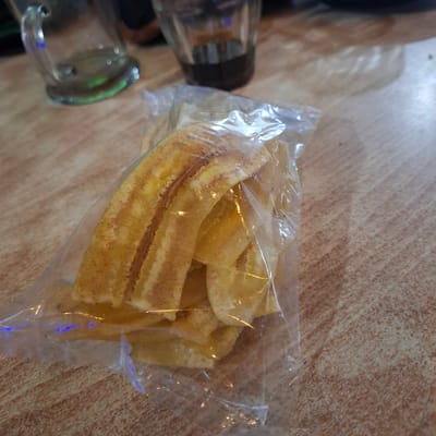 In Mesjid Tuha, Indonesia, a clear plastic bag filled with banana chips rests on a wooden table. The banana chips are thinly sliced and have a golden-yellow color with darker brown streaks, suggesting they are fried or caramelized. A small amount of seasoning, possibly cinnamon, appears to be sprinkled on them. In the background, out of focus, are two clear glass cups, one containing a dark liquid, likely coffee. The table's surface has a wood grain pattern.
