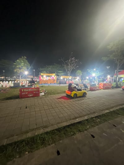 An exterior nighttime street scene in Sodong, Indonesia, captures an open-air food and entertainment market. In the foreground, a paved walkway is lined with grass and drainage grates. To the right, a bright yellow toy car with two occupants, a child and an adult, is illuminated by the scene's ambient lighting. The car is moving along the walkway, heading towards the right of the frame.
Beyond the walkway, a grassy area is populated with various stalls and attractions. A prominent red stall in the middle foreground displays the word "TAKOYAKI" in white lettering and appears to be a food vendor. To its left, a person is attending to a cooking station. Further back, several other stalls are visible, some with colorful banners and what appear to be games or merchandise displays. In the background, trees and streetlights dot the landscape, contributing to the overall illumination of the market. The sky is dark, indicating it is nighttime.