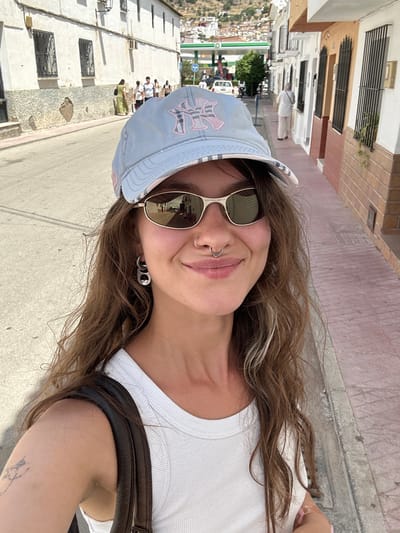 A young woman with long, wavy brown hair, wearing a light blue baseball cap with pink lettering and sunglasses, smiles at the camera. She has a septum piercing and large hoop earrings. She is wearing a white ribbed tank top and a dark brown shoulder bag strap is visible. She is standing on a paved street in what appears to be a town, with buildings lining both sides of the street. In the background, a white car is parked near a gas station with a green canopy. Several other people are walking in the distance, and a hilly, populated area is visible behind the buildings. The scene appears to be Pizarra, Spain, based on contextual information.