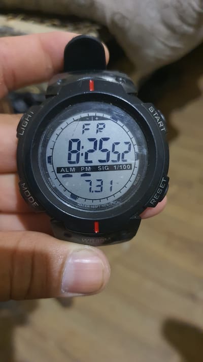 A close-up shot shows a digital watch face with black casing and a grey dial. The watch is held by a person's thumb and forefinger, with the thumb positioned at the top of the watch and the forefinger near the bottom. The watch face displays the time as 8:25:52, with "FR" above it, likely indicating Friday. Below the time, "ALM PM SIG 1/100" is visible, suggesting alarm, PM, signal, and a stopwatch function. The number "7.31" is displayed below that. The watch also has labels around the dial: "LIGHT" on the left, "START" on the top right, "RESET" on the bottom right, and "MODE" on the bottom left. The watch is also labeled "WATER 165FT RESIS" and "WR50M" indicating water resistance. The background is blurred, appearing to be a textured surface, possibly a couch or fabric, in muted tones. The scene is not identifiable as being in Karankul, Uzbekistan.