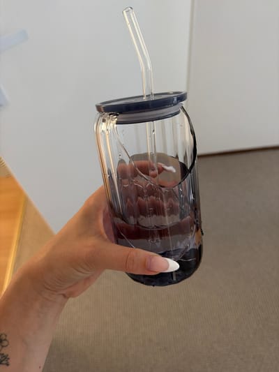 In Ridderkerk, Netherlands, a person's hand, with a flower tattoo on the wrist and manicured nails, holds a cracked glass tumbler filled with a dark liquid. A clear glass straw is inserted through the dark lid. The tumbler has vertical ridges and water droplets on its exterior. The background consists of a light-colored wall and a textured carpet.
