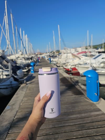 A person's hand, with painted fingernails and a tattoo on the forearm, holds a lilac-colored thermos cup in the foreground. The thermos has a white lid with a light-colored button and a black logo that reads "MEOKY". The background depicts a marina with numerous sailboats docked in the water under a clear blue sky. The boats are moored along a wooden pier, and the scene is likely Jagodje, Slovenia. Two blue bollards are visible on the pier, one in the middle ground and another further in the background. The overall atmosphere is sunny and maritime.