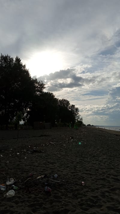 The image depicts a beach in Pasi ie Leubeue, Indonesia, with a dark, sandy shore littered with plastic waste. Tall, dark trees line the left side of the frame, partially obscuring structures made of what appears to be thatch or woven material. The sky is overcast with the sun peeking through, casting a bright glow in the upper left quadrant. In the distance, the ocean is visible with a few people walking along the shoreline. The foreground is dominated by the strewn trash, including plastic bottles and cups, indicating a significant pollution problem on the beach.