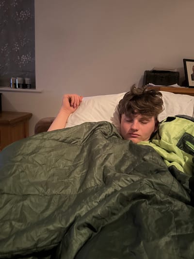 A young man with brown hair is sleeping on a bed with a green and lime green comforter. He is lying on his back with his left arm outstretched, his head resting on a white pillow. The scene is located in a bedroom in a house in the city of Newton upon Derwent, United Kingdom. The location is based on coordinates (latitude: 53.939, longitude: -0.906). The bedroom is lit by natural light coming from a window on the left. A wooden nightstand is visible on the left side of the image. The image is taken from the perspective of someone looking down at the man from the end of the bed.