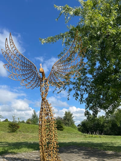 The image depicts a golden, openwork sculpture of a bird, possibly a phoenix, standing in a park-like setting. The sculpture is intricately detailed with swirling patterns forming its body and wings. It is positioned on a paved area, likely a pathway, with a grassy hill and trees in the background. The sky is a vibrant blue with scattered white clouds. To the right of the sculpture, a leafy green tree branches overhead. The overall impression is one of a bright, sunny day in an outdoor public space. The sculpture is the primary subject, a decorative element within a natural landscape. There are no people or animals visible in the image. The location is identified as Азярэцкі сельскі Савет, Belarus.