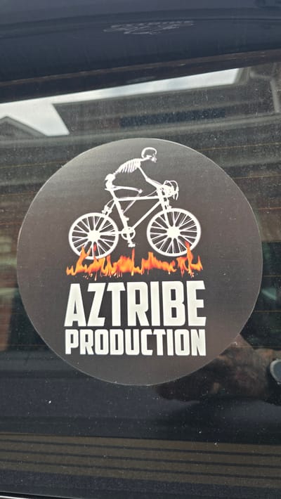 A circular sticker is adhered to the rear window of a vehicle. The sticker features a white skeleton riding a white bicycle with white spoked wheels. Flames in shades of orange and yellow are depicted beneath the bicycle's wheels. Below the flames, the text "AZTRIBE" is displayed in large, white, block letters, followed by "PRODUCTION" in slightly smaller, white, block letters. The sticker is dark gray with a white graphic and text. The background of the image is the interior of a vehicle, with the reflection of the exterior visible on the window. The sticker is located in Milton, Canada, on the rear window of a parked vehicle.