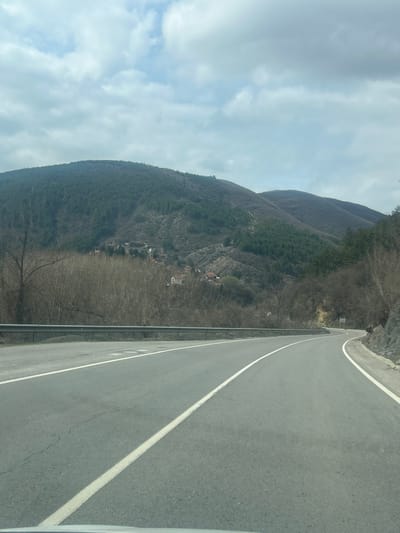 The image displays a two-lane asphalt road curving to the right through a mountainous region near Lukovo, Bulgaria, under an overcast sky. The road features continuous white lines marking both edges and a broken white line delineating the center. A silver metal guardrail parallels the left side of the road. To the left, a mountain slope is covered with bare deciduous trees in the foreground and lower sections, transitioning to dense coniferous forest higher up. A cluster of approximately six to eight houses with visible reddish-brown roofs is situated on the mid-slope of this mountain. The right side of the road features a rocky cut and bare deciduous trees, with a small, unreadable white road sign visible further down the curve. Distant mountains in the background are also partially forested with mixed deciduous and coniferous trees. No individuals, animals, or other vehicles are present in the visible scene. The perspective is from the interior of a vehicle, looking forward.