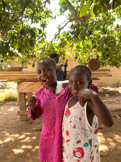 Two smiling young girls are positioned closely together, with the girl in front wearing a pink dress and the girl behind her in a white top decorated with strawberries. They are standing outdoors, with a leafy tree canopy above them and a dirt ground below. In the background, a wooden structure and a person holding a cell phone can be seen. The location is Hoss, Nigeria.