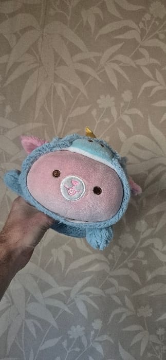 In Tompsan, Bulgaria, the image depicts an unidentified individual's hand holding a plush toy against a patterned wall. The plush toy features a pink, oval-shaped face with two small, dark brown circular eyes and a light pink embroidered snout design. It is wearing a light blue hooded costume, complete with small pink ears and a prominent golden horn, suggesting it represents a unicorn or similar mythical creature. The human hand, visible from the wrist down on the lower-left, has sparse hair and supports the toy from below. The background consists of a wall covered in muted beige or light brown wallpaper, adorned with a repeating pattern of lighter-toned, stylized botanical elements, including various leaves and small flowers.