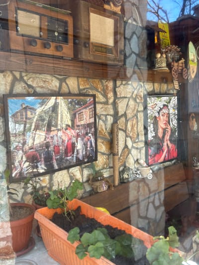 This image shows a window display in what appears to be a shop or establishment in Bukovlak, Bulgaria. The interior exhibits a collection of items, including framed pictures or artworks. One features a woman with floral elements, while another depicts a scene with people in what may be traditional Bulgarian clothing and architecture. A wooden bat with the word "GANESHA" inscribed on it is visible. An old-fashioned radio is also present. Additionally, the display incorporates potted plants in terracotta and orange containers. The entire arrangement is set against a stone-patterned wall or backdrop. The scene is indoors, observed through a window.
