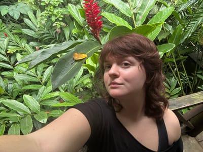 An image depicts a Caucasian female subject positioned in a natural, lush green environment consistent with tropical gardens found in areas like Flores, Guatemala. The female, with medium-length, wavy reddish-brown hair and a neutral expression, looks directly at the camera, appearing to take a selfie. She wears a black top, possibly off-the-shoulder, with a visible strap on her right shoulder. Her left arm is partially visible in the lower-left foreground. The background is dominated by dense, vibrant green foliage comprising various broad-leafed tropical plants. A prominent red ginger flower (likely *Alpinia purpurata*) is visible in the upper center-right, rising above the other plants. On a large green leaf to the left of the red flower, a single yellow, withered leaf rests horizontally. In the lower right corner, a section of a weathered, dark wooden structure, possibly a railing or a bench, is partially visible behind the subject.