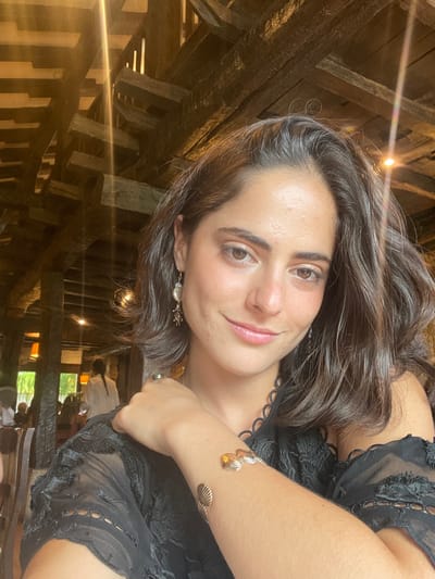 An adult female is depicted in a portrait-style photograph taken inside Restaurante Zelaieta in Amorebieta-Etxano, Spain. The location is identifiable by its distinctive interior architecture, featuring a dense network of heavy, dark wooden beams forming the ceiling and structural elements of the dining area. The female subject, positioned in the foreground, has medium-brown skin, dark brown, wavy hair, and brown eyes, and is looking directly at the camera with a subtle smile. She is wearing a dark, possibly black, semi-sheer garment with patterned sleeves. Accessories include drop earrings with a pearl-like component on her left ear, a ring on her right ring finger, and a gold-toned cuff bracelet adorned with multiple shell-shaped motifs on her right forearm. Her right hand is visible, resting near her left shoulder. In the blurred background, other patrons are seated at tables in the dimly lit environment, indicating a public dining setting.