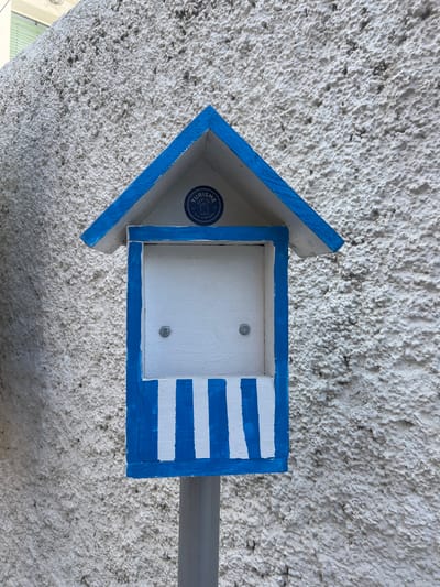 A blue and white arrow-shaped directional sign is prominently featured, pointing towards the left against a heavily textured, off-white wall. The sign appears to be made of painted wood, exhibiting a distinct nautical or Mediterranean aesthetic.
The arrow's body is a rectangular box with blue borders and a white interior. On the far right side of this box, a blue section features three horizontal white stripes, evoking the design of a flag. Inside the central white panel of the box, two metallic screws or bolts are visible. The arrow's head, also outlined in blue, contains a circular blue sticker with white text. Partially legible words on the sticker include "TURISME," "SANT POL DE MAR," "CASA," and "VILA," along with an emblem resembling a stylized house or building. This text indicates the sign's function as a tourism guide or marker for the location Sant Pol De Mar.
The sign is attached to a grey, rectangular support structure that extends horizontally to the right. The background wall has a coarse, stucco-like finish, showing numerous small bumps and scattered dark specks, possibly dirt or mold. The lighting suggests it is daytime. The image is captured from a slightly low angle, looking up at the sign mounted on the wall.