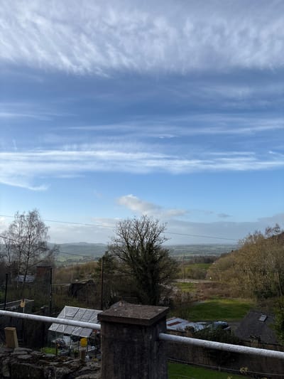 This image depicts an outdoor panoramic view of a rural landscape in Snailbeach, United Kingdom, captured from an elevated perspective. The sky occupies the upper two-thirds of the frame, featuring a clear blue expanse with multiple layers of wispy cirrus and altocumulus clouds, casting a bright, daylight illumination.
In the foreground, a stone wall, likely part of a retaining wall or balcony, is visible at the bottom of the frame, topped with mossy stones and partially obscured by foliage. A thick, white-painted metal railing or pipe runs horizontally across the lower mid-section of the image, attached to a large, grey, square stone pillar on the left.
The midground reveals a sloped terrain characterized by mixed vegetation. To the left, several bare deciduous trees stand with intricate branch patterns. A white-framed glass greenhouse is prominently positioned below the railing, containing various garden items, including a yellow bucket and potted plants. Further back, glimpses of brick structures, potentially parts of houses or outbuildings, are visible. Overhead, multiple power lines traverse the scene horizontally.
Towards the right midground, more deciduous trees, some still bare and some with nascent foliage, line a downward slope. A dark-colored SUV is partially visible, parked near a building with a dark-tiled roof featuring a skylight. A patch of green grass is present in this area.
The background presents a vast, rolling landscape of green fields and distant hills under a hazy sky, indicating a significant depth of field. No individuals or active interactions are observed within the scene. The overall impression is one of a quiet, rural village setting on a clear day, possibly in late winter or early spring due to the bare trees.