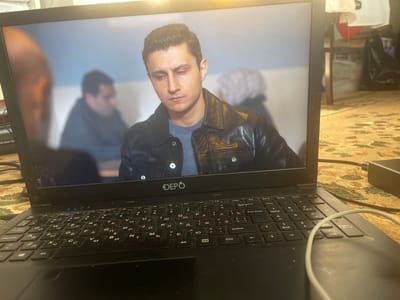 The image shows a laptop displaying a scene from a media program. The main subject on the screen is a man with dark hair, wearing a black leather jacket over a grey t-shirt. He has a somber expression and is looking downwards. In the background on the screen, out of focus, are two other figures. One is on the left, with their profile visible and a reddish-brown complexion. The other is in the middle, appearing to be a man with short dark hair, also blurred.
The laptop itself is black and has a visible keyboard with Cyrillic characters. The brand name "DEPO" is visible on the bezel below the screen. The laptop is placed on a surface covered with a patterned rug or carpet, which has predominantly beige and red tones. The surrounding environment appears to be indoors, with some clutter visible in the background to the right of the laptop, including what looks like a table, some white fabric hanging, and black trash bags. The lighting suggests it could be daytime, as there is a bright light source visible above the laptop screen. The overall impression is of a casual viewing setting.