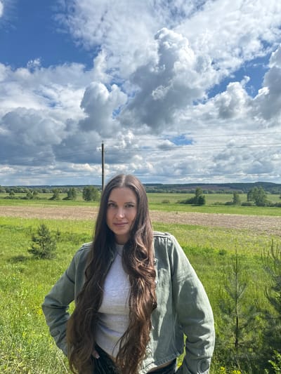 The image shows a young woman with long brown hair, wearing a grey shirt and a green jacket, standing in a field in Kremyonki, Russia. The background features a landscape with a mix of green fields, cultivated land, scattered trees, and a distant treeline. The sky is predominantly blue with large, puffy white clouds. A telephone pole is visible.