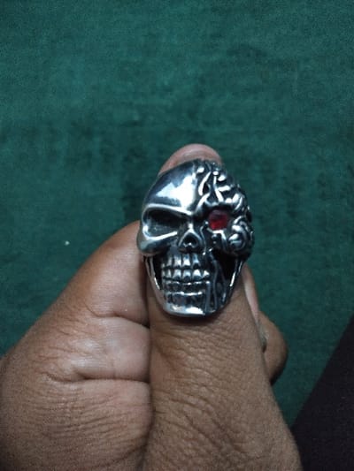 The image displays a close-up of a silver-colored ring worn on a person's thumb. The ring is designed as a skull, featuring intricate details. The right side of the skull (viewer's left) depicts bone structure, while the left side (viewer's right) is stylized to expose brain-like convolutions. A single red gem is set into the left eye socket (viewer's right) of the skull. The skull design includes visible teeth. The thumb, positioned vertically in the center of the frame, exhibits a medium-to-dark brown skin tone, showing creases and texture. The remaining fingers are partially visible at the lower left. The background consists of a soft-focus, dark teal or deep green textured surface, likely fabric, which provides no identifiable environmental details or specific geographical context within Ciudad de San José, Costa Rica. The scene is lit from the front, accentuating the metallic surface of the ring.