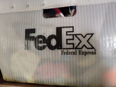 The image displays a close-up, angled view of a white, ribbed plastic bin with the "FedEx Federal Express" logo printed on its side. The logo is in black and consists of the letters "FedEx" in a distinctive font, followed by "Federal Express" in a smaller, serif font, and a registered trademark symbol. The bin appears to be translucent, with various items, some of which are brightly colored fabrics, visible through it. The bin is resting on a wooden surface. There is a rectangular cutout at the top of the bin, through which more of the contents are visible. The overall condition of the bin suggests it is used, with some smudges and discoloration. The scene does not contain any elements that would suggest a specific location in the city of Gal’chino, Russia.