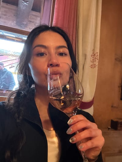 A female subject is depicted indoors, looking directly at the camera, with her lips touching the rim of a wine glass. She has dark hair and is wearing a dark zip-up jacket over a light-colored top. Her right hand, adorned with light-colored nail polish, holds the clear stemmed glass which contains a pale rosé-colored liquid, approximately half full. The glass is etched with "HOSPIZ ALM ST. CHRISTOPH" and a logo. In the upper left background, through a window, a partial view of a blurred male figure in a dark jacket is visible against an outdoor, snowy environment with a red structure. A wooden beam is visible above the window. To the right, a red curtain and a beige curtain with red embroidered patterns hang against a light brown wall. The location is Hospiz Alm in St. Christoph, Austria.