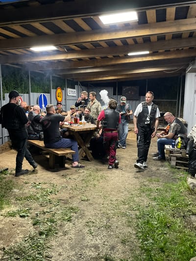 A group of individuals, likely bikers, are gathered at a picnic table under a wooden structure in Doma 45 Kilometr, Russia. The scene appears to be outdoors at night. Some individuals are seated at the picnic table, while others are standing or sitting on wooden pallets nearby. Some of the individuals are drinking. One person is vaping or smoking, as evidenced by the white smoke. Several road signs are visible near the table, including one with a speed limit of 40 km/h and a "STOP" sign. The environment consists of dirt, grass, and a metal fence. Some of the individuals are wearing biker vests. One person is wearing black boots and pants. The area is lit by fluorescent lights attached to the wooden structure above.