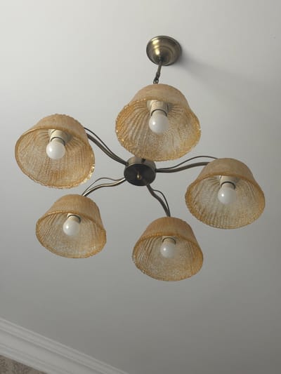 A ceiling-mounted chandelier with five amber, textured glass shades hangs from a white ceiling. Each shade encloses a white, globe-shaped light bulb. The chandelier has a central brass fixture with curved arms extending to support the shades. The image appears to be taken from below, looking up at the chandelier. The location is the village of Samakhvalavichy, Belarus.