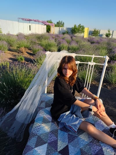 A young woman with shoulder-length brown hair and bangs is sitting on a quilted blanket spread over a bed frame in a lavender field. She is wearing a black t-shirt and light blue shorts, and a white watch on her left wrist. She has bracelets on her right wrist. A sheer white veil drapes over the bed frame and extends to her left. The background is a vast field of purple lavender under a clear blue sky. A white corrugated metal fence runs along the horizon, with trees and a yellow door visible in the distance. The sun is low in the sky, casting a warm glow and long shadows, suggesting it is late afternoon or early evening. The overall mood is serene and peaceful. There is no visible text in the image.