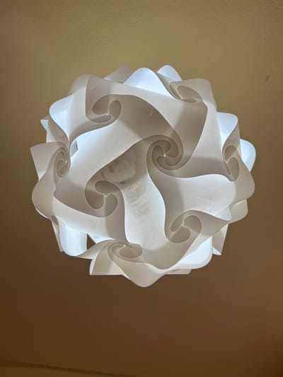 The image displays an illuminated, spherical modular lamp shade constructed from interlocking translucent white plastic pieces. The lamp, emitting a bright white light, is suspended from a light brown or beige ceiling in an interior space located in Vilanova I La Geltrú, Spain. The individual plastic components are curved, creating a complex, layered, and swirling pattern across the lamp's surface. The background ceiling is out of focus, indicating the lamp is the primary subject. No individuals, actions, or additional objects are visible within the frame.