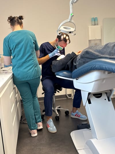 The image depicts a dental treatment room in Bretteville-sur-Odon, France. In the foreground, a male dentist, clad in dark blue scrubs, a light blue surgical mask, and blue gloves, is seated on a rolling stool, leaning over a supine patient. He wears a headlamp and holds a dental instrument with an integrated light, focused on the patient's mouth. His feet are visible, wearing light blue sneakers with red stripes. To the left, a female dental assistant, viewed from the back, stands at a white counter. She wears teal scrubs and has her hair styled in two buns. On the counter, a computer keyboard is visible, alongside various items. The patient, partially visible, is lying on a blue and white dental chair, wearing dark pants and a dark top. An articulating overhead dental light is positioned above the patient. The room features white walls, a dark grey floor, and includes a white wall-mounted paper towel dispenser and two stacked white cups on a shelf in the background.