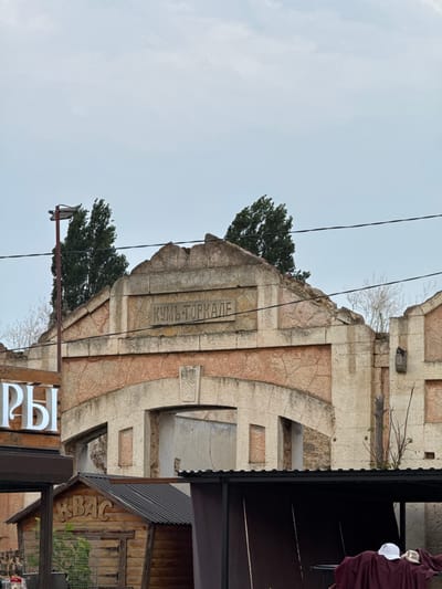 The image shows the entrance to an abandoned building with the Cyrillic inscription "КУМЬ-ТОРКАЛЕ" above an arched opening. The building appears to be made of stone and has a weathered, dilapidated facade. In front of the building, there is a small wooden structure with the Cyrillic inscription "КВАС" on its roof, suggesting it might be a vendor stall. A neon sign with Cyrillic letters that reads "РЫ" is partially visible on the left side of the image. The sky is overcast, and some trees are visible behind the building. The overall scene depicts a rundown, possibly historic, location. The image is captured from a slightly low angle, looking up at the entrance.