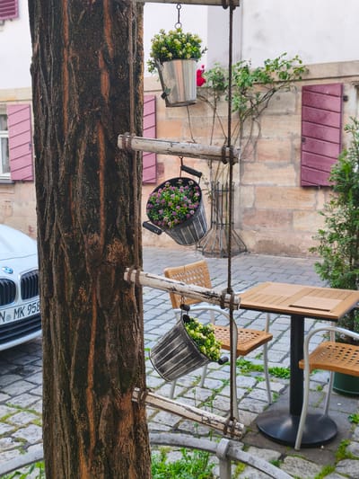 The image depicts an outdoor seating area, likely a cafe or restaurant, in Erlangen, Germany. A large tree trunk is prominent in the foreground, with a rustic ladder-like structure attached to it. Several metal buckets filled with greenery and small purple flowers are suspended from this structure by ropes, adding a decorative element.
In the background, a building with a light-colored stone facade and closed maroon shutters is visible. A white car with a German license plate, reading "N MK 958", is parked to the left of the tree. Further to the right, a small outdoor table with two chairs is set up on a cobblestone patio. The overall scene suggests a pleasant, possibly quiet, moment in a European town.
The lighting indicates it is daytime, with soft light suggesting it could be morning or late afternoon. There is no visible human activity. The mood is tranquil and inviting, with the plants adding a touch of natural beauty to the urban setting. The presence of the cafe furniture and the car hint at a functional, lived-in space within the town.