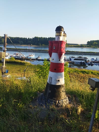 An eye-level, medium shot captures a decorative lighthouse situated on a grassy embankment overlooking a calm river in Leverkusen, Germany. The lighthouse, painted in alternating red and white horizontal bands with a black base and top, features a decorative ladder on its side. Behind it, the river is dotted with various boats, including motorboats and larger houseboats docked at the water's edge. The opposite bank of the river is lined with a dense forest, and the sky above is a clear, pale blue. To the left of the lighthouse, a metal pole with electrical boxes and wires is visible, indicating infrastructure near the riverfront. The foreground is dominated by tall, dry grass illuminated by the warm glow of the setting sun.