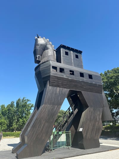 The image depicts a large, dark wooden structure resembling the Trojan Horse, set against a clear blue sky. The structure is intricately constructed with visible wooden planks and has multiple openings that appear to be windows. The horse's head is visible on the left side, with a detailed wooden design. The setting includes trees and some paved ground, suggesting an outdoor location. A green metal fence is visible on the right side of the structure. There are no people clearly visible in the image, but a small sign with text is attached to the horse's side. The sunlight suggests it is daytime, and the clear sky indicates fair weather. This structure is a replica and is located in Çanakkale Merkez, Turkey, likely a tourist attraction related to the legend of the Trojan War. The overall impression is one of historical representation and an outdoor exhibition.