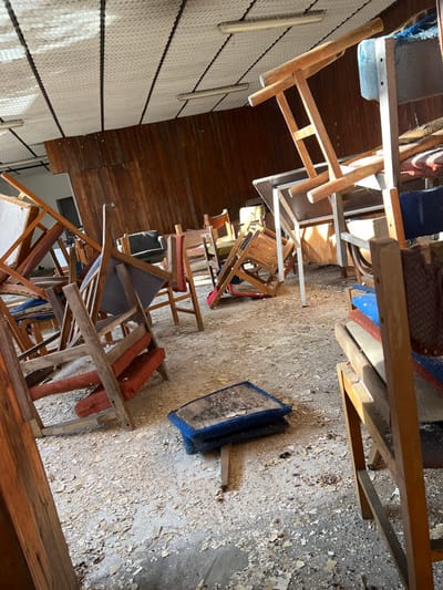 The image portrays an interior scene within a building in Stoianovo, Bulgaria, characterized by extensive disarray. The concrete floor is largely covered with light-colored debris, consistent with flaked paint or plaster. Numerous wooden chairs, estimated at over two dozen, are scattered throughout the space, many overturned, stacked haphazardly, or leaning against other furniture. These chairs exhibit fabric upholstery in various states of wear, featuring colors such as red, blue, green, and brown. Two tables are partially visible: one with a light-colored top and metal legs, positioned in an overturned and stacked configuration, and another with a wooden top and wooden legs, also in disarray. In the immediate foreground, two blue, soiled chair cushions lie detached on the floor. The rear wall of the room is covered with dark, reddish-brown wooden paneling. The ceiling consists of white, possibly textured acoustic tiles or insulation panels, intersected by dark structural beams, with two non-illuminated fluorescent light fixtures attached. Natural light enters from the left side and overhead, creating distinct shadows across the scene. No individuals are present in the depicted area.