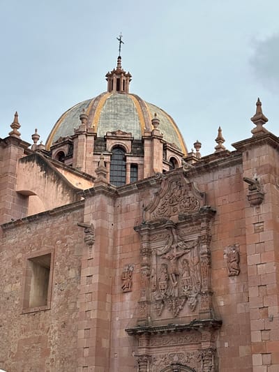 The image shows a close-up, low-angle view of the pink stone facade of the Zacatecas Cathedral in Zacatecas City, Mexico. The cathedral's prominent dome, partially obscured by the surrounding structure, is visible at the top, topped with a cross. Intricate stone carvings, including a crucifixion scene and religious figures, adorn the facade. Decorative elements like spires, gargoyles, and arched windows are also present. The sky is overcast, casting a soft light on the building.